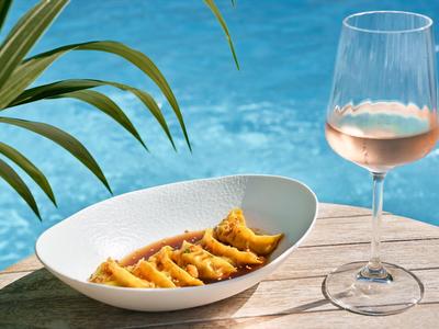 Wein in einem Glas und Snacks auf einem Tisch neben einem Pool und Palmenblättern.