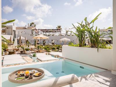 Sunny hotel pool area with white buildings, sun loungers, umbrellas, and a floating tray with breakfast.
