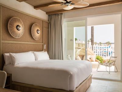 Schlafzimmer mit großem Bett, hellem Holz, Wanddeko und Balkon mit Blick auf Stadt bei Sonnenschein.