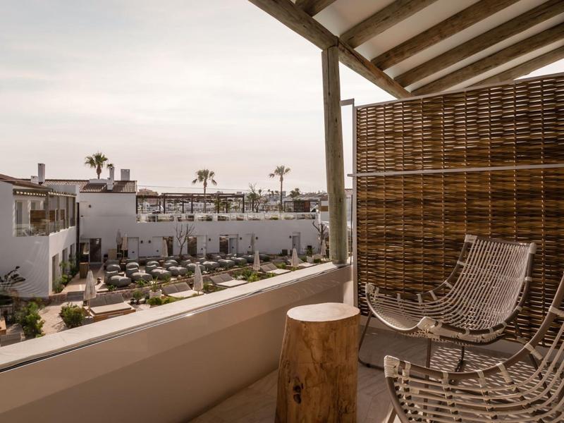 Balcony with wicker chairs overlooks a modern hotel pool area with palm trees under cloudy sky.
