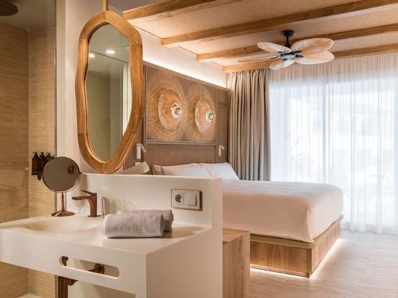 Bright hotel room with wooden decor, large bed, mirror, and sheer curtains lighting the space.