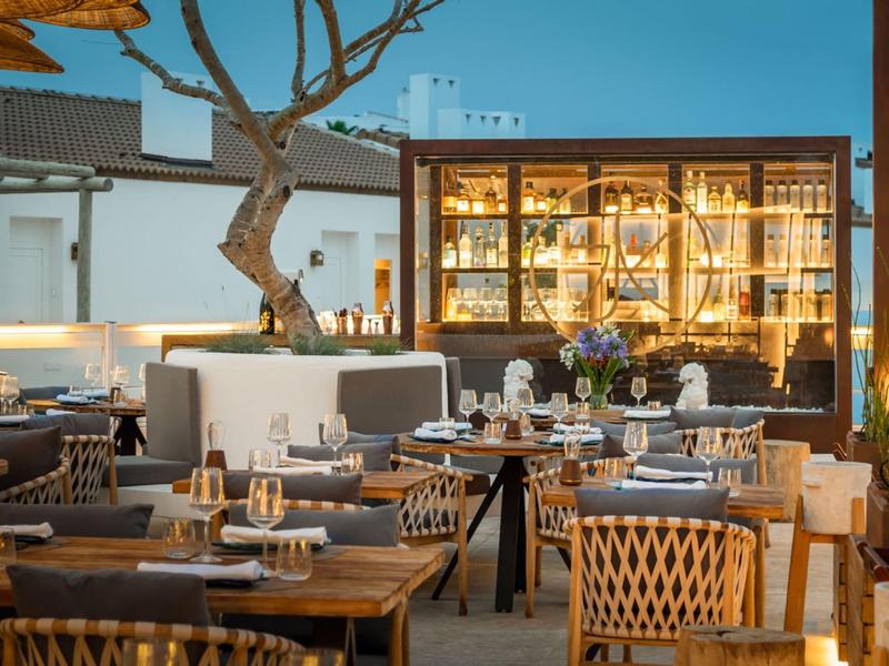 Cozy outdoor restaurant dining area with wooden tables, cushioned chairs, and warm lighting at dusk.