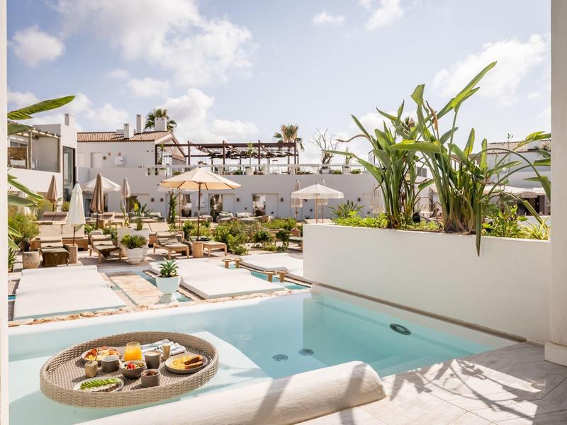 Sunny hotel pool area with white buildings, sun loungers, umbrellas, and a floating tray with breakfast.