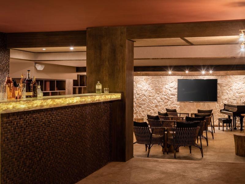 Cozy hotel lounge with backlit stone bar and dark wooden tables and chairs under warm lighting.