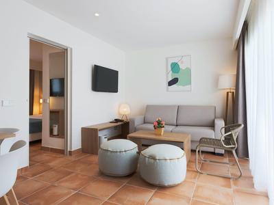 Lichte woonkamer met bank, salontafel, twee poefs, tv en zicht op aangrenzende kamer.