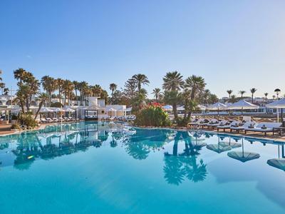 Groot buitenzwembad met palmbomen, ligstoelen en een heldere blauwe lucht op de achtergrond.