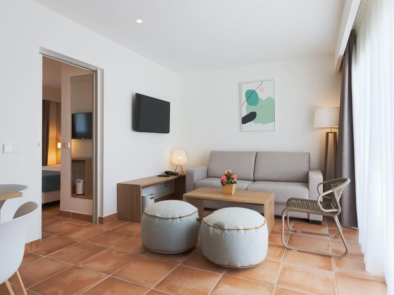 Lichte woonkamer met bank, salontafel, twee poefs, tv en zicht op aangrenzende kamer.