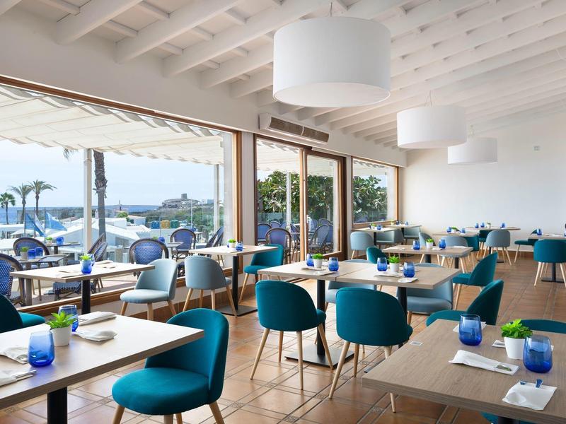 Modern restaurant met grote ramen, blauwe stoelen en uitzicht op zee bij zonnig weer.
