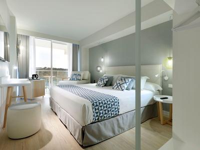 Nowoczesny pokój hotelowy z dużym łóżkiem, biurkiem, fotelem i dużym jasnym oknem.