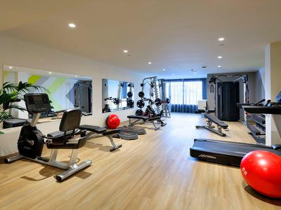 Jasna siłownia z drewnianą podłogą, różnym sprzętem treningowym i dużymi oknami.