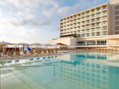 Basen na zewnątrz z leżakami i dużym nowoczesnym hotelem pod częściowo zachmurzonym niebem.