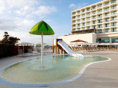 Dziecięcy basen z zjeżdżalnią i zielonym parasolem obok nowoczesnego hotelu.