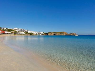 Plaża z jasnym piaskiem, przezroczysta woda, biały hotel na skalistym wybrzeżu pod błękitnym niebem.