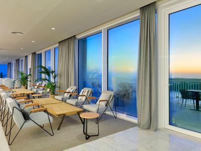 Nowoczesna sala z dużymi oknami i stołami, widok na taras o zachodzie słońca.