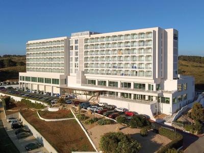 Duży nowoczesny hotel z balkonami, otoczony ogrodami i parkingami pod jasnym, niebieskim niebem.