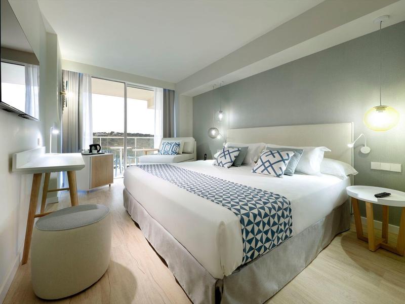 Nowoczesny pokój hotelowy z dużym łóżkiem, naturalnym światłem i minimalistycznym wyposażeniem.