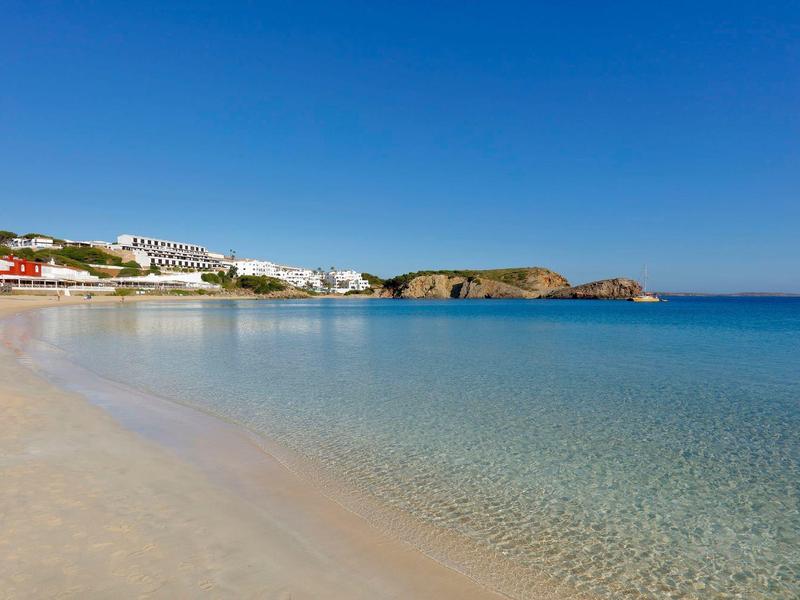 Plaża z jasnym piaskiem, przezroczysta woda, biały hotel na skalistym wybrzeżu pod błękitnym niebem.