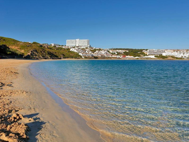 Piaszczysta plaża z czystą wodą i białym hotelem w tle pod niebieskim niebem.