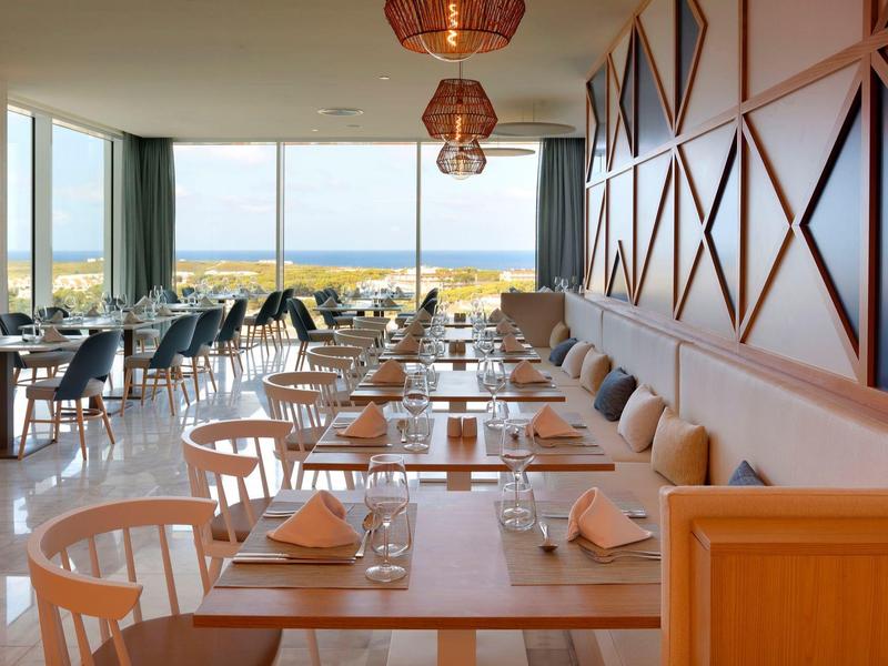 Jasna restauracja z dużymi oknami, widokiem na morze, drewnianymi stołami i nowoczesnym wystrojem.