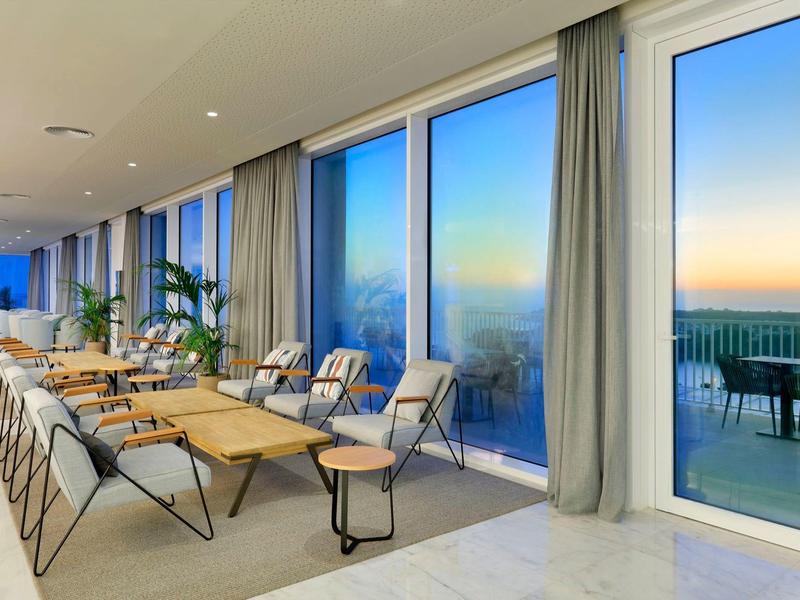 Nowoczesna sala z dużymi oknami i stołami, widok na taras o zachodzie słońca.