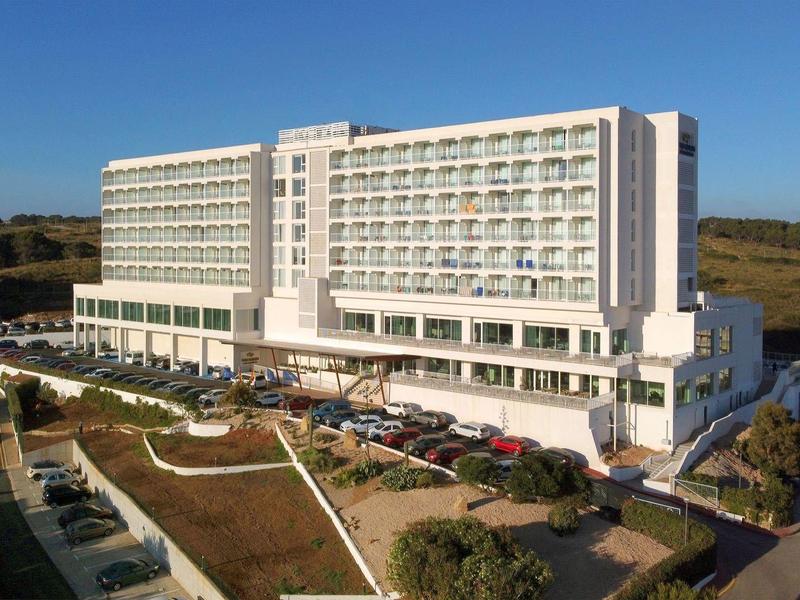 Duży nowoczesny hotel z balkonami, otoczony ogrodami i parkingami pod jasnym, niebieskim niebem.