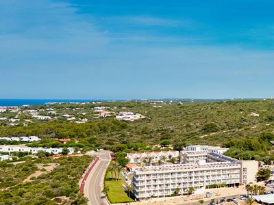 Vista di un hotel in un paesaggio verde con cielo azzurro e mare sullo sfondo.