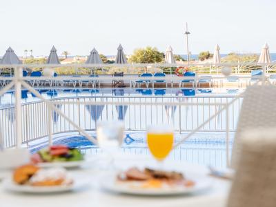 Vista di una piscina con ombrelloni, davanti un tavolo con due piatti e un bicchiere di succo d'arancia.
