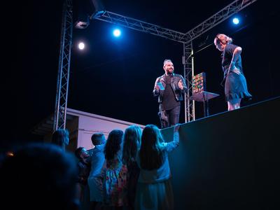 Due persone parlano su un palco illuminato davanti a un pubblico di notte.