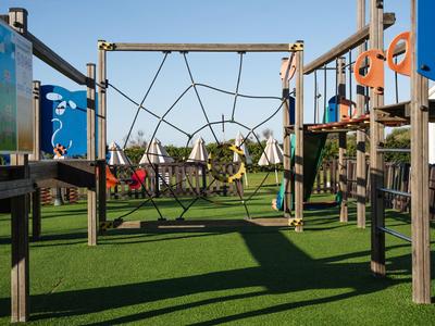 Parco giochi con rete da arrampicata e scivolo su prato sintetico verde sotto un cielo sereno