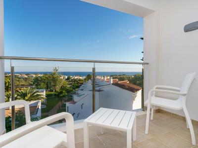 Balcone con sedie bianche e tavolo, vista sul mare e sul giardino sotto un cielo azzurro.