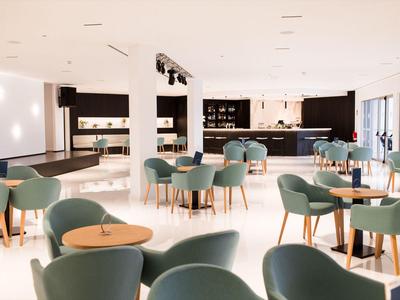 Hall d'hôtel moderne et lumineuse avec fauteuils verts et tables en bois sur sol clair.