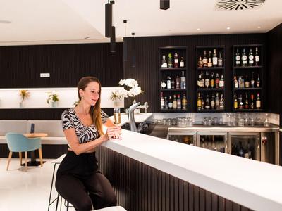 Femme assise au bar d'un hôtel moderne avec des étagères remplies de bouteilles en arrière-plan.