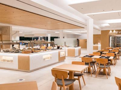 Restaurant d'hôtel lumineux avec chaises en bois, tables et buffet proposant divers plats.
