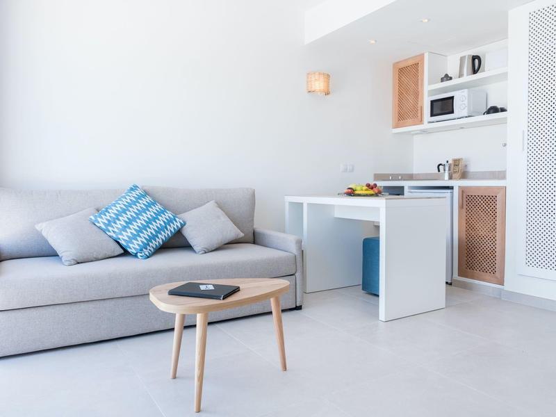 Sala de estar moderna y luminosa con sofá gris, cojín azul, pequeña mesa de madera y cocina abierta.