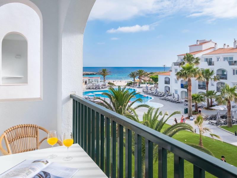Balcón con silla, mesa y jugo de naranja con vista al mar y edificios blancos bajo cielo azul.