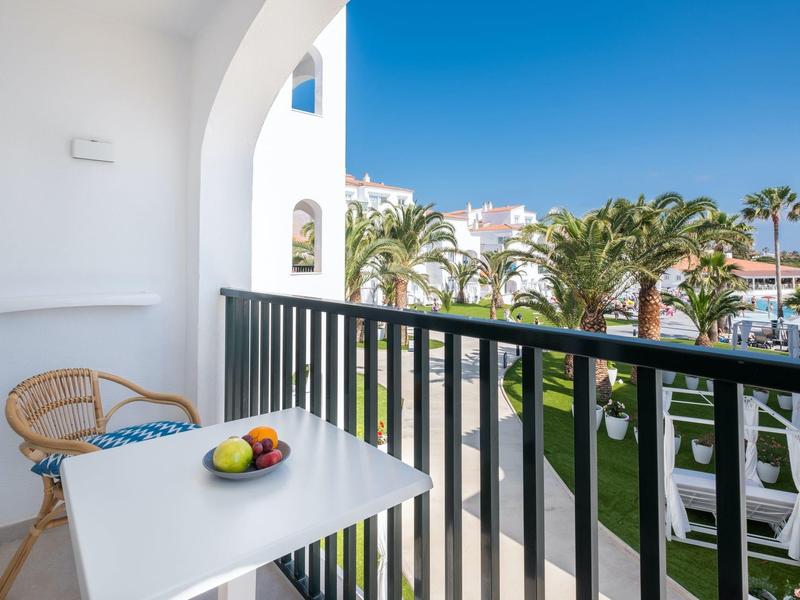 Balcón con mesa, silla y vista al jardín con palmeras y cielo azul.