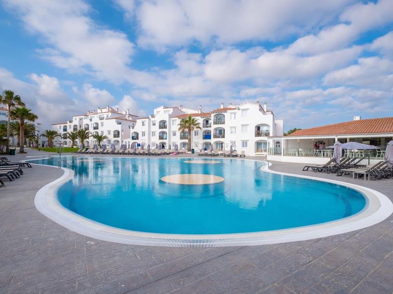Gran piscina de hotel con tumbonas y edificios blancos bajo cielo azul.