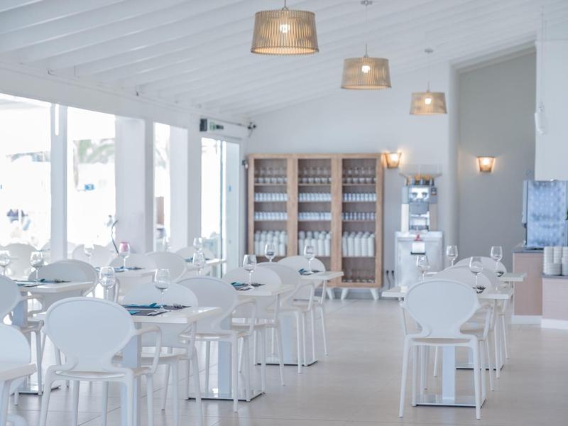 Restaurante moderno y luminoso con mesas y sillas blancas y grandes ventanas para luz natural.