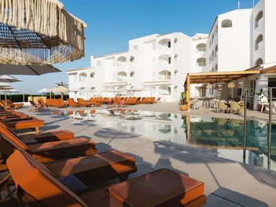 Espace piscine d'hôtel moderne avec transats, parasols et bâtiments blancs sous ciel clair.