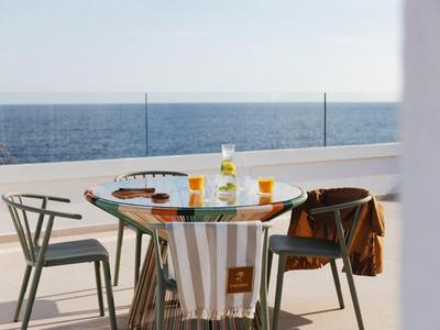 Table en verre moderne sur terrasse avec vue sur la mer, boissons et trois chaises.