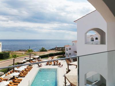 Vue depuis un balcon sur une piscine et la mer avec des bâtiments blancs et des chaises longues.
