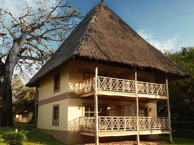Traditionelles zweistöckiges Holzhaus mit Strohdach und Balkon, umgeben von grüner Wiese und Bäumen.