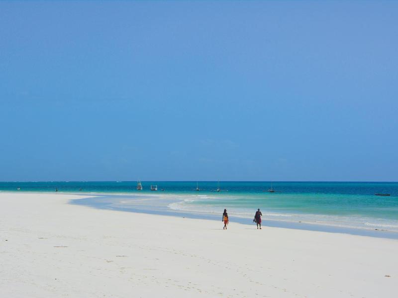Weißer Sandstrand mit blauem Himmel und zwei Personen am Ufer.