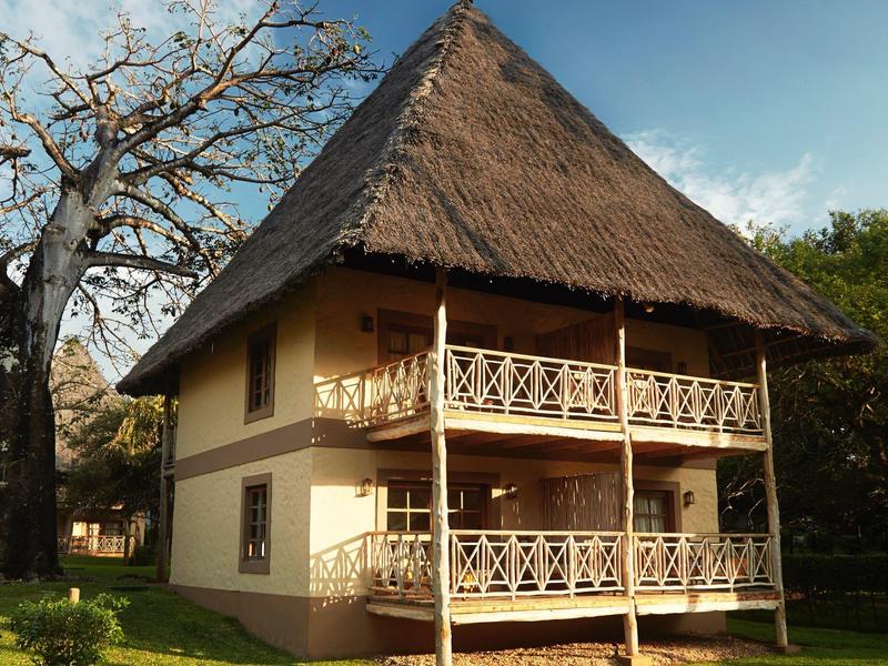 Zweistöckiges Haus mit Strohdach und zwei Balkonen, umgeben von Bäumen und grünem Rasen.