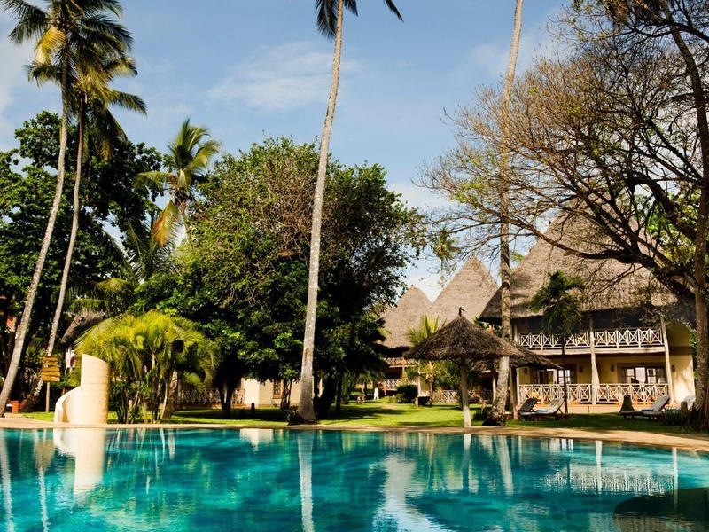 Ein klarer blauer Pool vor einem Haus mit vielen Bäumen und Palmen an einem sonnigen Tag.