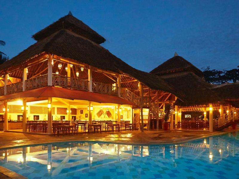 Beleuchtetes, traditionelles Holzhaus neben einem hellblau beleuchteten Swimmingpool bei Nacht.