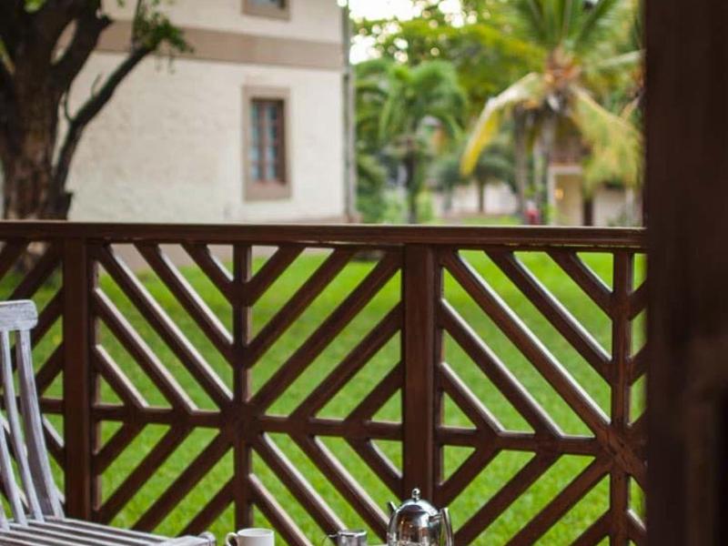 Balkon mit Holztisch, Teekanne, Tassen, zwei Stühlen, grüner Garten und Häuser im Hintergrund.