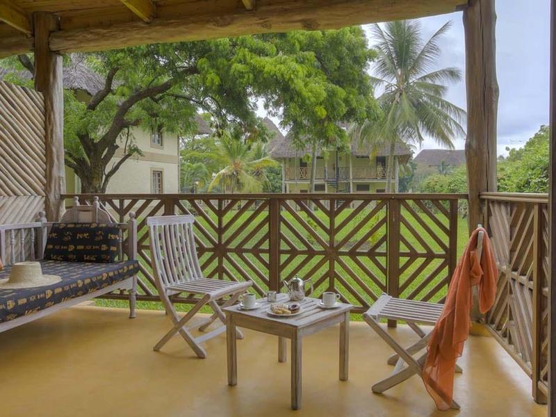 Überdachter Balkon mit Holzgeländer, zwei Stühlen, Tisch und grüner Pflanzenkulisse im Hintergrund.