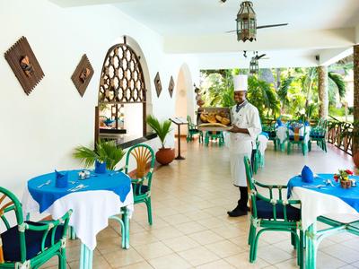 Area ristorante coperta con tavoli e sedie, cameriere che serve bevande, area esterna verde visibile.