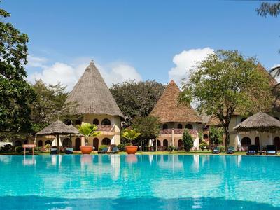 Großer blauer Pool vor runden, strohgedeckten Häusern und Bäumen unter blauem Himmel.